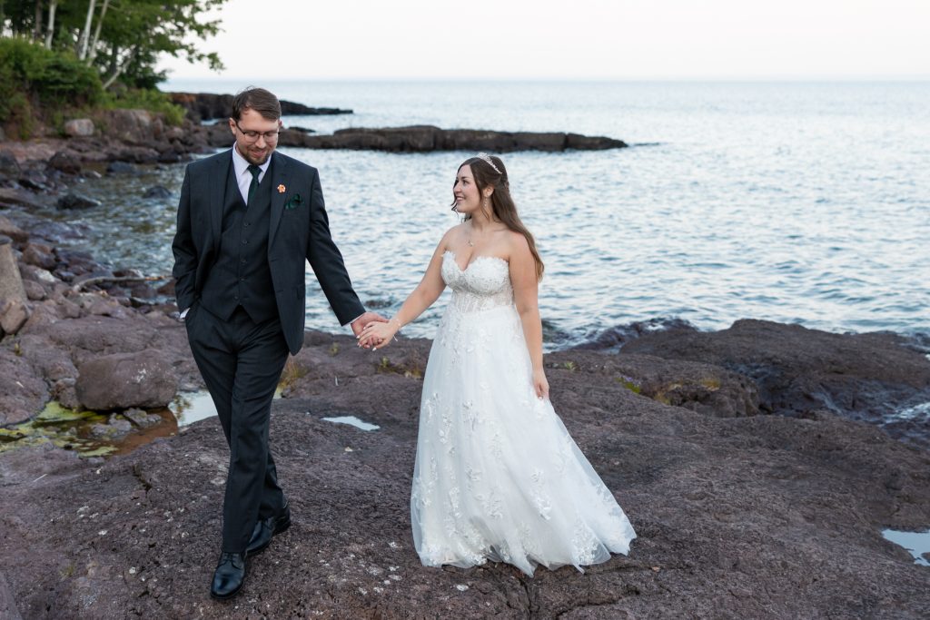 Newly married couple holding hands and walking along a rocky shoreline, showcasing wedding photography services