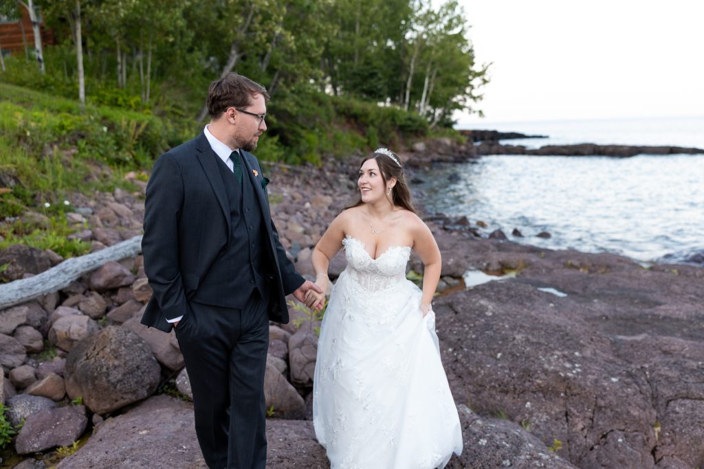 Newly married couple holding hands and walking along a rocky shoreline in Wisconsin, showcasing