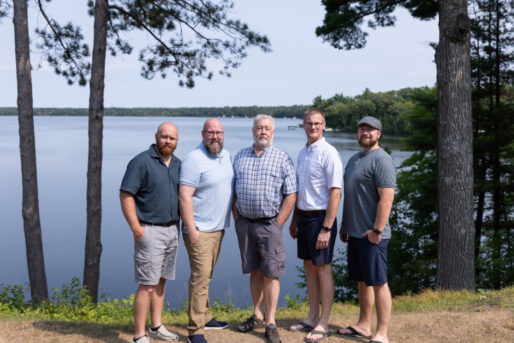 Five men of varying ages pose for a family portrait near a serene lake
