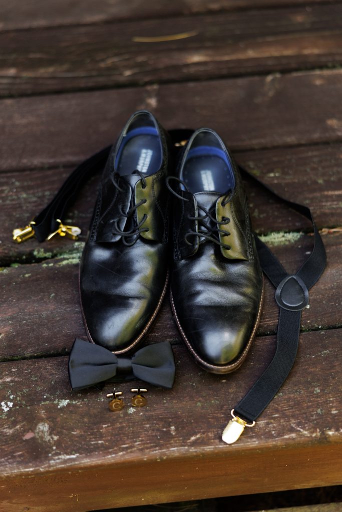 Elegant black leather dress shoes, suspenders, bow tie, and cufflinks arranged for a wedding detail