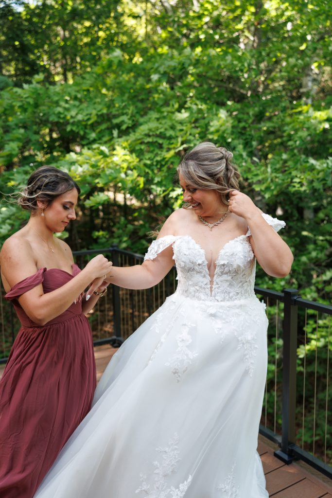 Bride in a detailed white wedding gown smiles as her bridesmaid assists with earrings