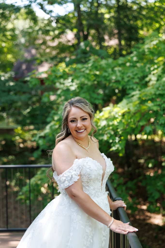 Bride Bianca in a detailed off-the-shoulder wedding gown smiles radiantly amidst lush greenery at Lakewoods