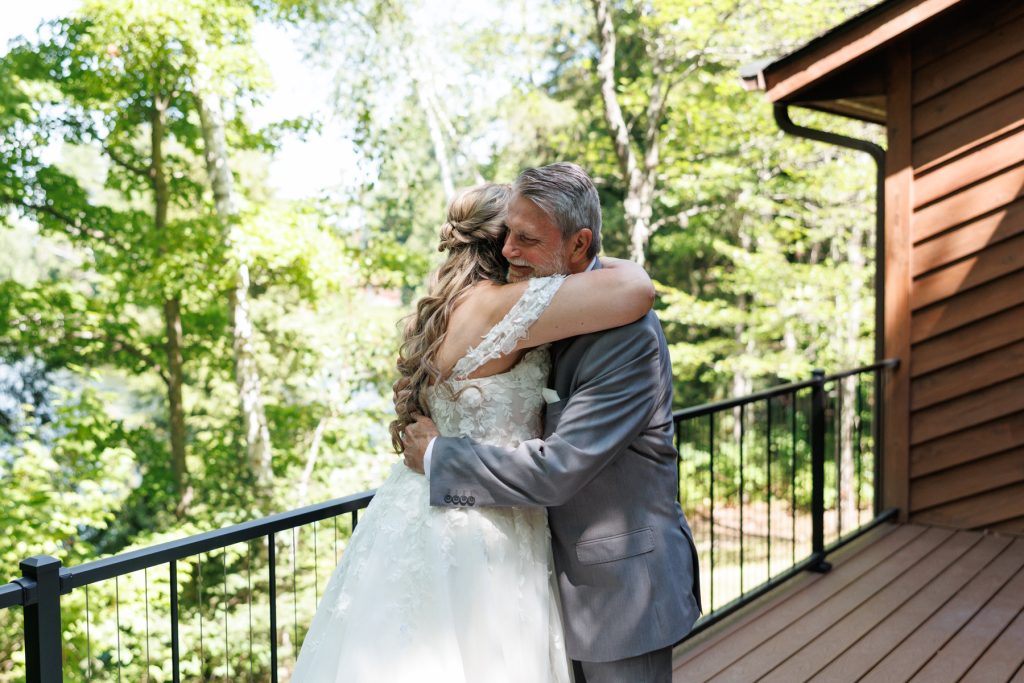 Bride in a lace wedding dress hugs her father on a wooden deck