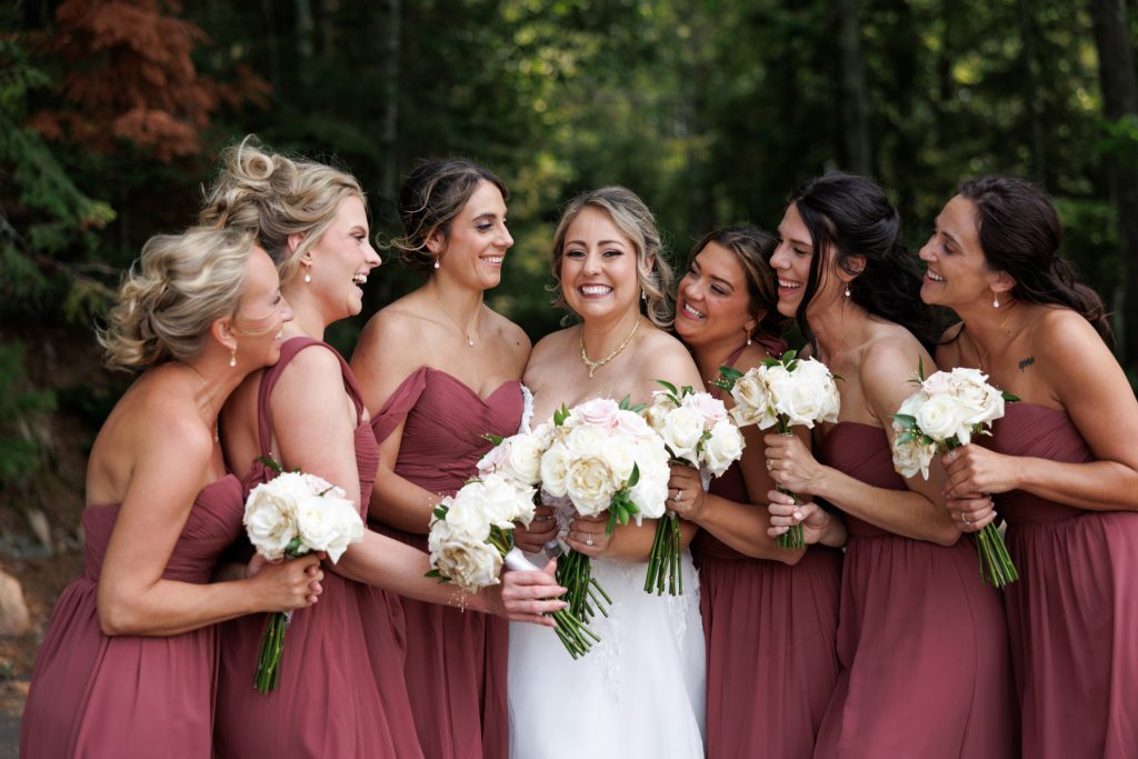 Bride and bridesmaids in burgundy dresses smiling and laughing together, holding bouquets of white roses