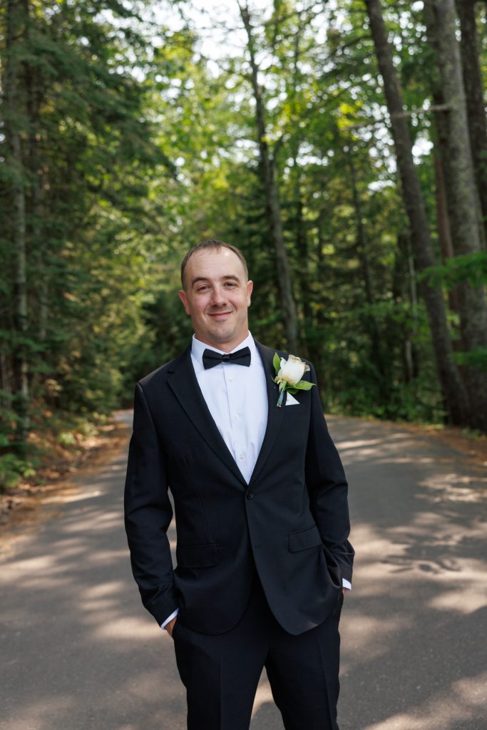 Groom in a black suit and bow tie smiles confidently on a wooded road