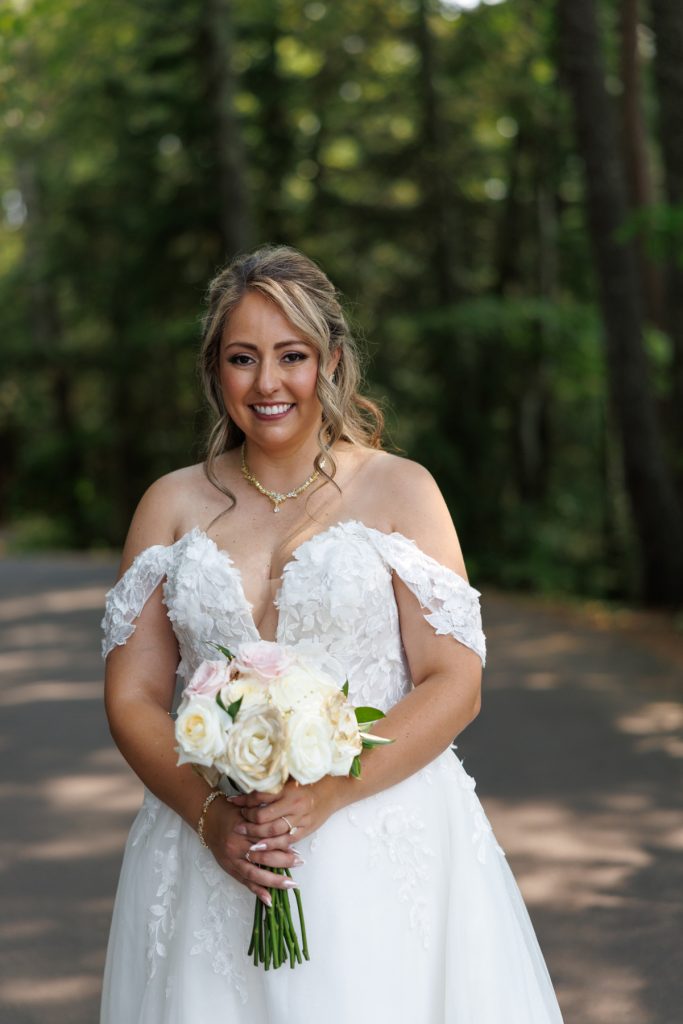 Bride Bianca in an off-the-shoulder wedding gown smiles and holds a bouquet of white roses