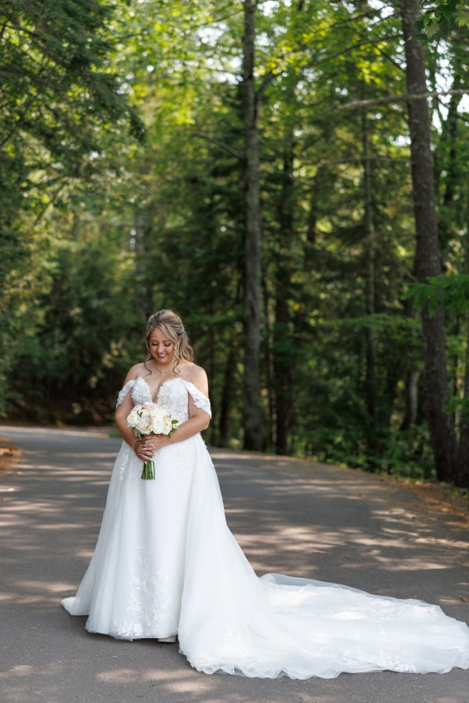 Bride Bianca in a flowing white wedding gown stands on a lakeside road at Lakewoods