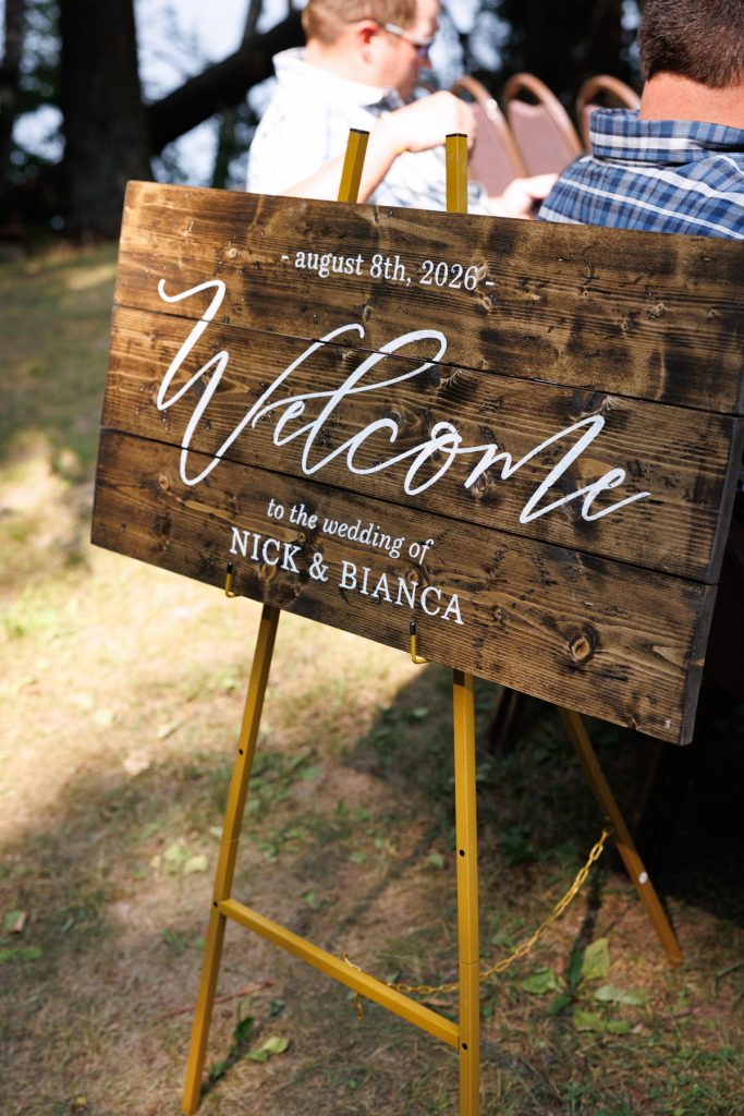 Rustic wooden welcome sign displaying "Welcome to the wedding of Nick & Bianca, August 8th