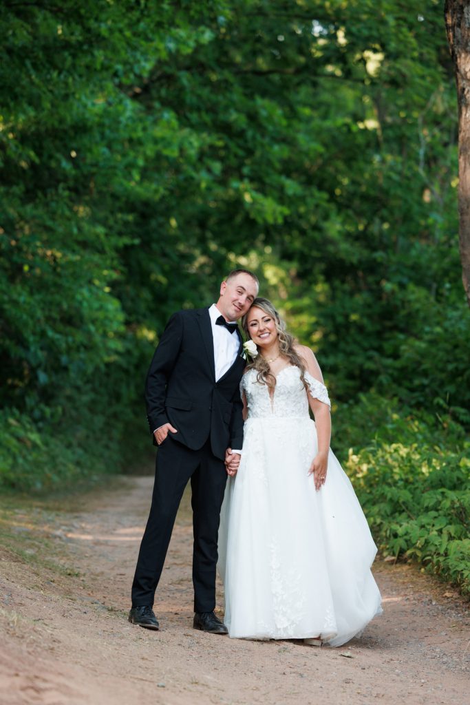 Bianca and Nick, the newlyweds, stand hand-in-hand on a wooded path at their Lakewoods Resort