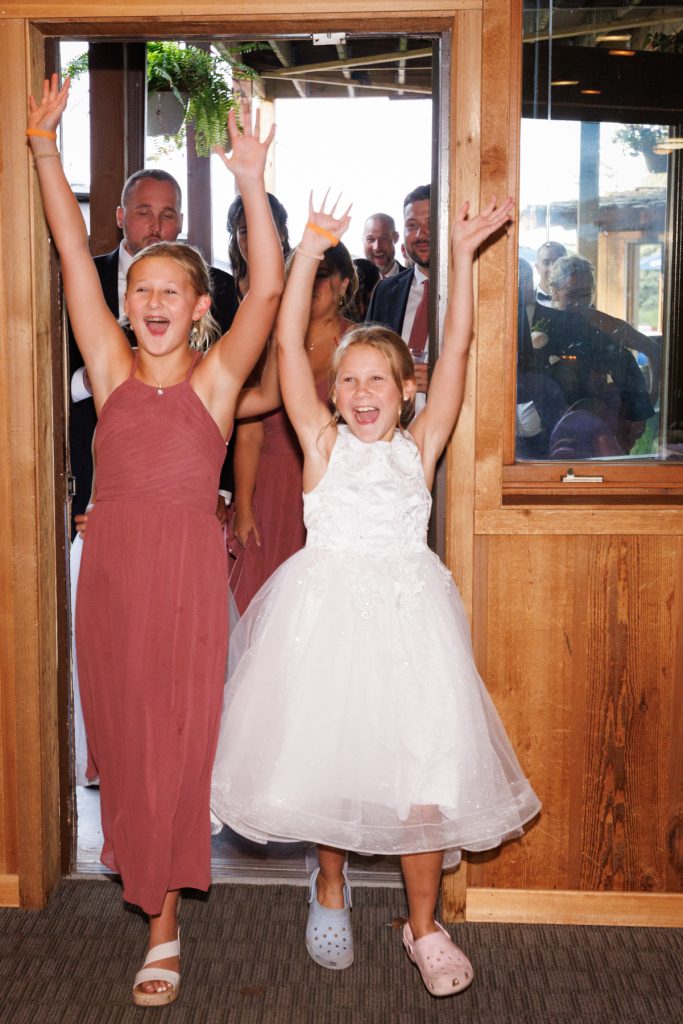 Two young flower girls with arms raised in excitement walking through a doorway
