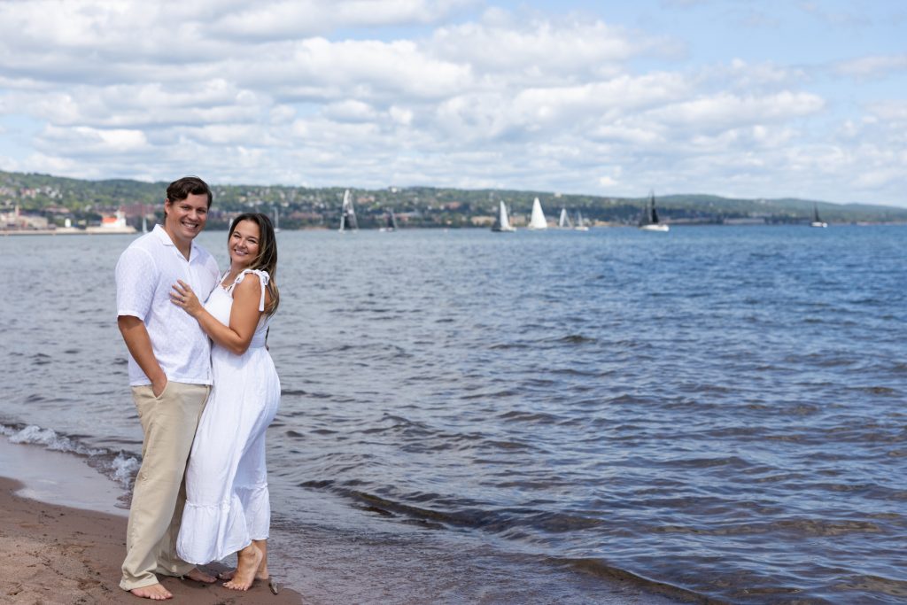 Cassie and Daniel, the engaged couple, stand on a sandy beach with Lake Superior
