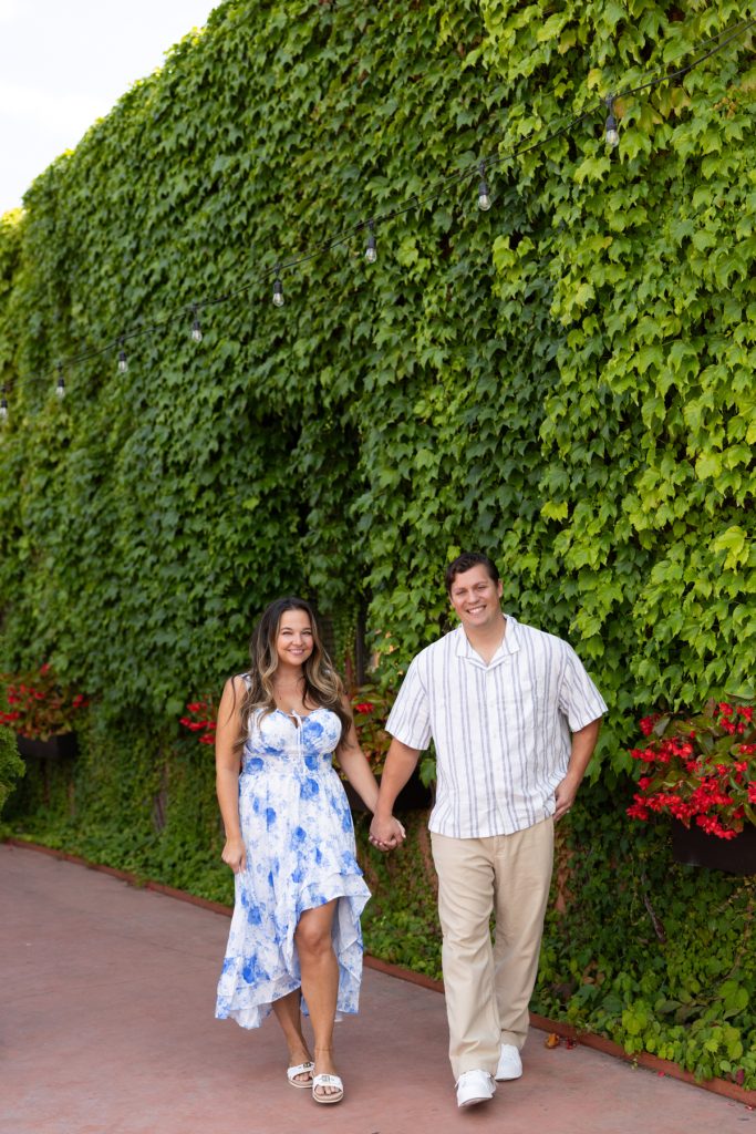 Cassie and Daniel, the engaged couple, walk hand-in-hand against a lush green wall of ivy