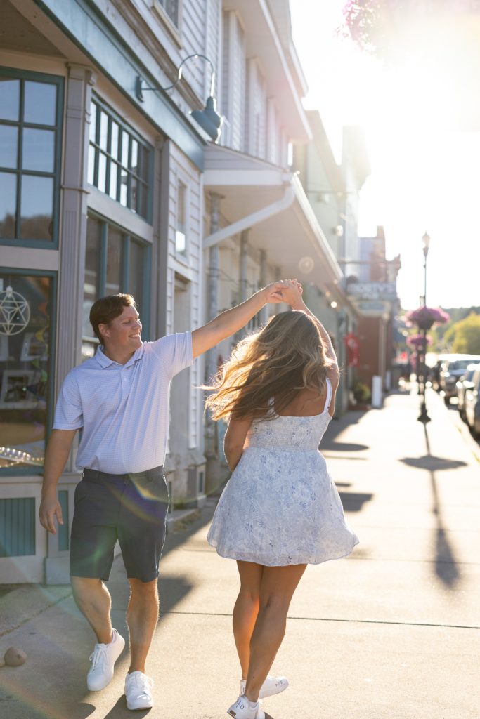 Cassie and Daniel, the engaged couple, joyfully dance together on a sunny street in Bayfield