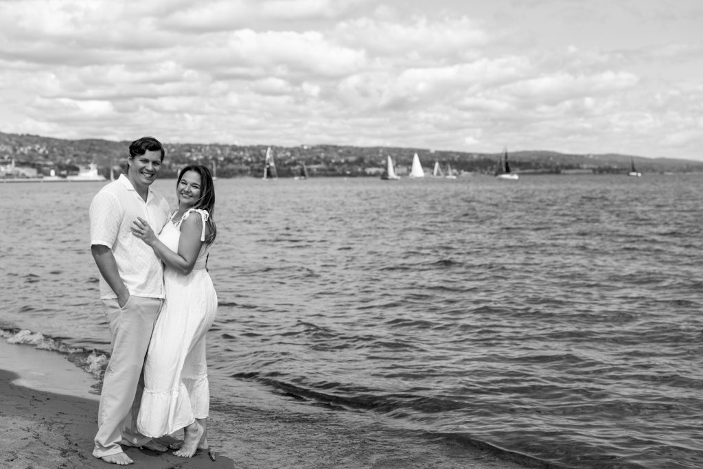 Cassie and Daniel, the engaged couple, stand on a Lake Superior shoreline with sailboats visible